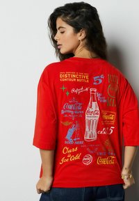 Czerwona, oversize'owa koszulka z vintage grafiką Coca-Coli w kolorach niebieskim, żółtym i zielonym, z półdługimi rękawami i luźnym krojem.