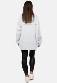 Witte textuur oversized shirt met verlaagde schouders, gecombineerd met glanzende zwarte leggings en witte sneakers. Uitzicht van achter.