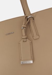 Borsa a tracolla sintetica beige con texture liscia, caratterizzata da un'etichetta in hardware argento e dal logo "Calvin Klein" impresso. Include un dettaglio con cinturino ad anello.