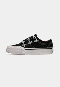 Zapatillas de lona negras con herrajes plateados, que cuentan con una lengua acolchada, suela de goma blanca y punta de tiro estriada. Costuras en contraste a los lados.