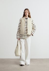 Γυναίκα με κρεμ επενδυμένο γιλέκο πάνω από μαύρο-λευκό ριγέ μακρυμάνικο πουκάμισο, λευκό φαρδύ παντελόνι, κρατώντας μια κρεμ τσάντα, όρθια μπροστά σε έναν λευκό τοίχο.