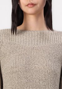 Femme avec des cheveux raides et foncés portant un pull en tricot beige à col montant côtelé, vue de la poitrine vers le haut.