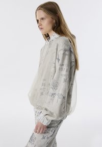 Sweatshirt léger gris avec un revêtement semi-transparent. Il présente des motifs imprimés et une capuche, avec des poignets et un ourlet côtelés.