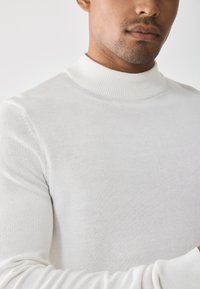 Suéter blanco de punto con un alto cuello de canalé. La tela es suave y lisa, con un diseño ajustado que se adapta al cuerpo.