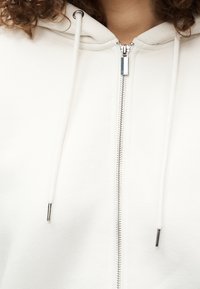 Λευκό φούτερ με φερμουάρ, κατασκευασμένο από απαλό βαμβάκι, με ψηλό γιακά, κορδόνια και ασημένιο φερμουάρ με μεταλλικές λεπτομέρειες.