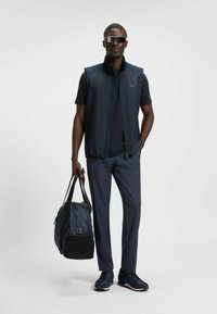 Marineblaues ärmelloses Weste über einem dunklen Hemd, kombiniert mit dunklen Hosen und Sneakers. Das Model hält eine dunkelblaue und schwarze Sporttasche.