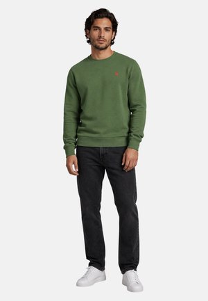 Mann trägt grünes Sweatshirt mit Rundhalsausschnitt, schwarze Jeans und weiße Turnschuhe, steht vor einfarbigem Hintergrund.