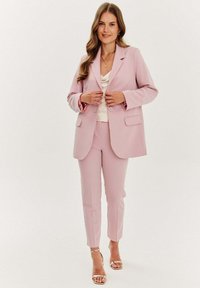 Světle růžový oblek skládající se z upraveného blejzru se dvěma kapsami a odpovídajícími slim-fit kalhotami. Pod ním je saténová bílá blůza.