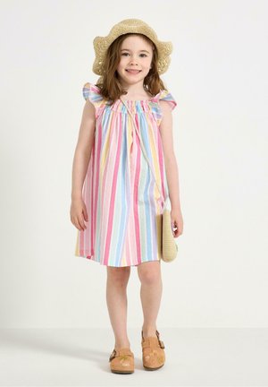 Ung pige smilende, iført en pastelstribet kjole, stråhat, brune sko med blomsterbroderi og holder en lille flettet taske.