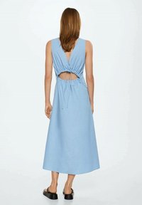 Eine Frau trägt ein ärmelloses, hellblaues Midikleid mit einem Rückenschlitz und Bindeband, kombiniert mit schwarzen Plateausandalen.