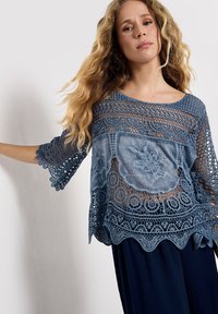 Top corto azzurro chiaro con intricate lavorazioni in pizzo, una texture trasparente e orlo a scallop. Maniche a 3/4 con dettagli in crochet aperto.