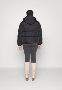 Giacca a vento nera con cappuccio, caratterizzata da un design trapuntato e una consistenza morbida. Indossata con jeans grigi e stivali beige fino al ginocchio.