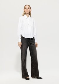 Woman standing wearing a white button-up shirt, black wide-leg jeans, and black pointed flats against a plain light background.