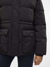 Črna podložena jakna z visokim ovratnikom, z dvema sprednjima žepoma, z zatičnimi gumbi in prešito teksturo. Nošena čez temen denim.