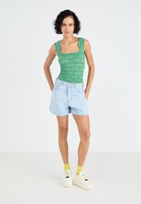 Grünes, strukturiertes, eng anliegendes Tanktop mit quadratischem Ausschnitt, kombiniert mit hellblauen Denim-Shorts. Dazu weiße Sneaker und gelbe Socken.