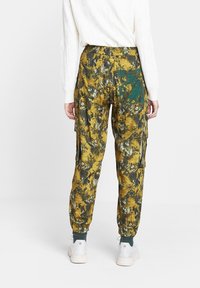 Bloemmotief cargobroek in mosterdgeel met groene accenten, voorzien van een elastische tailleband, zijzakken en taps toelopende boorden.