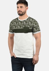 Camiseta de manga corta con un patrón de hojas de palmera en verde y blanco, un bolsillo en el pecho de color verde sólido y una sección inferior blanca. Material de algodón.