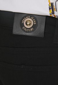 Černé džíny s koženou páskovou nášivkou a zlatým kováním. Nálepka nese nápis "VERSACE JEANS COUTURE" vytištěný a s raženými detaily.