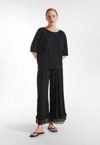 Musta, ylisuurikokoinen t-paita, jossa on lyhyet hihat, yhdistetty leveälahkeisiin mustiin housuihin, joissa on läpikuultava helma. Päällä mustat varvassandaalit.