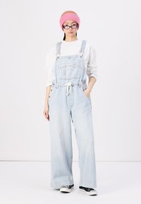Salopette en denim bleu clair avec des jambes larges, des bretelles réglables et des poches avant. Portée par-dessus un haut blanc, assortie avec des chaussures noir et blanc.