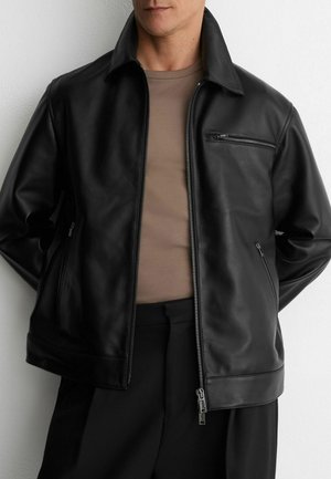 Mann trägt eine teilweise geschlossene schwarze Lederjacke über einem hellbraunen Hemd und schwarzen Hosen, steht vor einer weißen Wand.