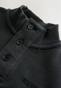 Tamsiai pilkas džemperis su austinėm apykakle, trimis juodais mygtukais ir tekstūruotu audiniu; ant apykaklės yra iškilniame rašte suformuotas logotipas.