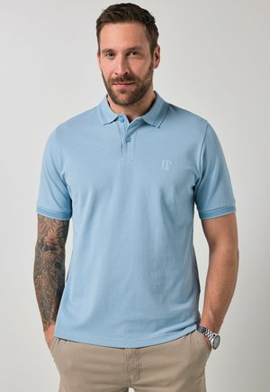 Mężczyzna z brodą, ubrany w jasnoniebieską koszulkę polo i beżowe spodnie, z tatuażem na lewej ręce i zegarkiem na prawej ręce, stojący na jednolitym tle.