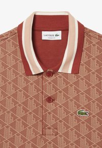 Röd mönstrad polotröja med krage som har krämfärgade ränder. Två röda knappar och en liten grön krokodil-logotyp på bröstet. Klassisk passform.