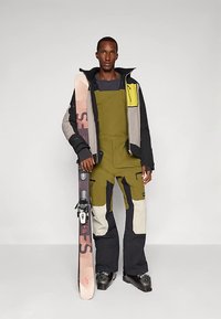 Mees, kellel on seljas roheline, must ja beež suusariietus, hoiab käes roosa ja musta suuski, seistes tasase helehall tausta ees.