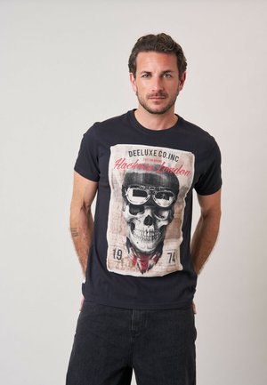 Man met kort bruin haar die een zwart T-shirt met een schedelafbeelding draagt, een veiligheidsbril en de tekst "Hackney London", staand tegen een effen achtergrond.