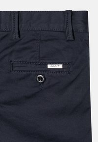 Marinblå bomullsbyxor med en framficka och knäppning, med en GANT-logotyp broderad på bakfickan. Slät textur.