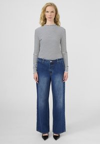 Žena s dugom plavom kosom nosi crno-bijelu prugastu majicu s dugim rukavima, široke plave traperice i crne cipele s oštrim vrhom.