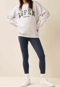 Felpa grigia con la scritta "ASPEN COLORADO" in lettere verdi scuro, abbinata a leggings blu scuro. Sneakers bianche con suole testurizzate e calzini a coste.