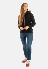 Felpa nera con zip e cappuccio, dotata di tasche frontali, abbinata a jeans blu e sneakers rosse con lacci bianchi e suola in gomma.