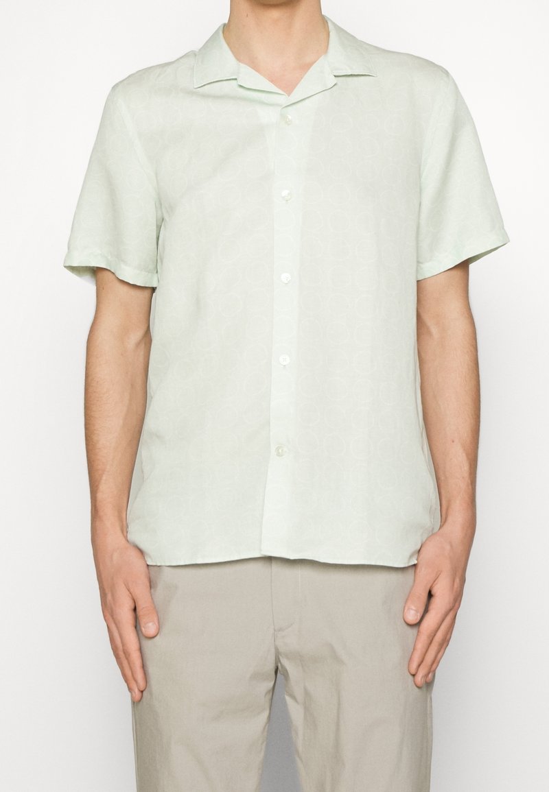 Homme portant une chemise à manches courtes de couleur vert pâle avec un motif circulaire subtil et un pantalon beige, debout devant un fond blanc uni.