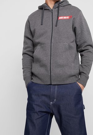 Homme portant un sweat à capuche gris zippé avec le logo « JUST DO IT. » sur la poitrine et un jean bleu foncé avec coutures blanches, mains dans les poches.