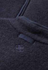 Navyblauwe wolmix stof met een zachte, gestructureerde oppervlakte. Heeft een slank ontwerp met een gestikt bloemenlogo en een stoffen lussticker.