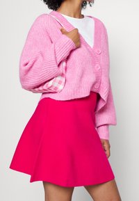 Cárdigan de punto rosa con textura acanalada y tres botones, combinado con una falda de tela brillante rosa con vuelo. Llevando un bolso de cuadros rosas.