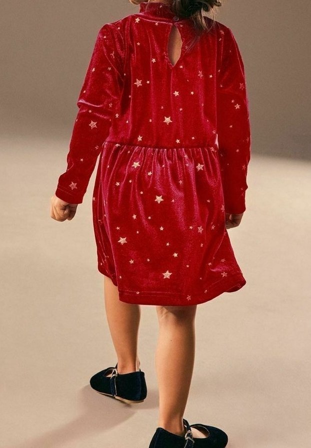 Enfant portant une robe en velours rouge avec des motifs d'étoiles, marchant de dos, assortie à des chaussures noires sur un fond uni.