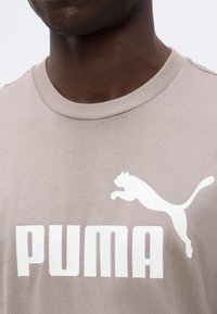 Gros plan d'une personne portant un t-shirt Puma beige avec un logo Puma blanc et un texte sur la poitrine.