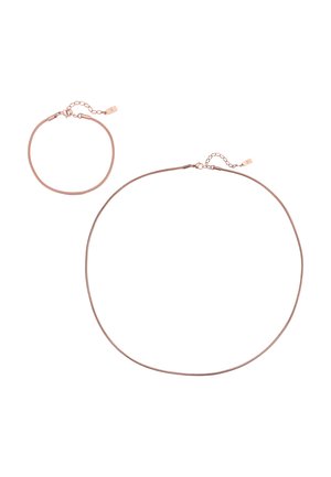 Mann trägt weißes ärmelloses Rippstrickshirt, dünne goldene Halskette und kleine Creolen, mit gepflegtem Bart und neutralem Hintergrund.