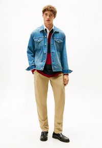 Chaqueta de mezclilla azul, con dos bolsillos en el pecho. Debajo, una camisa a rayas rojas y marino. Pantalones cargo beige y zapatos negros completan el atuendo.