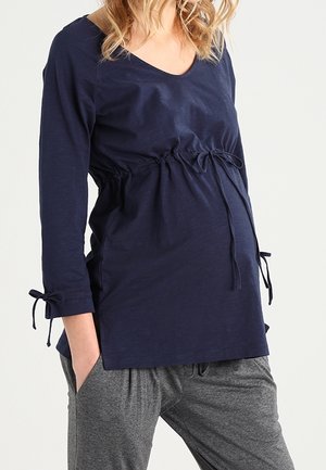 Kobieta w ciąży w granatowej bluzce ciążowej z regulowanymi sznurkami i szarych casualowych spodniach, stojąca z jedną ręką w kieszeni.