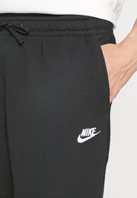 Pantalon de survêtement noir Nike avec taille à cordon et logo blanc brodé sur la cuisse gauche, porté avec une main dans la poche.