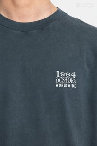 Ciemnoszara bawełniana koszulka z okrągłym dekoltem oraz białym nadrukiem z logo „1994 DC Shoes Worldwide” na lewej stronie klatki piersiowej.