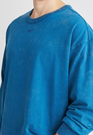 Person iført en falmet blå Nike sweatshirt med lange ærmer og et lille mørkt Nike-logo centreret under halsudskæringen.