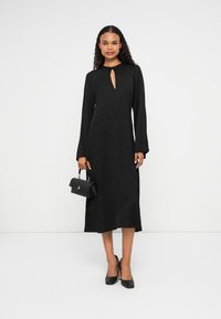 Vestido preto de mangas longas com decote em forma de buraco de chave, textura suave e silhueta ajustada. Combinado com uma pequena bolsa preta e saltos altos.