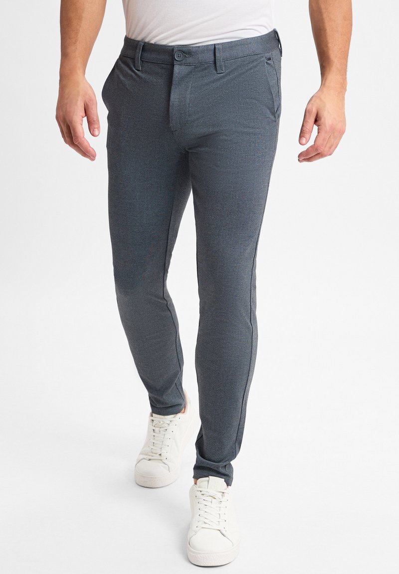 Pantalones ajustados de corte slim, grises y con un patrón en un tejido elástico texturizado, con frente plano, trabillas para cinturón y bolsillos laterales; combinados con zapatillas blancas.