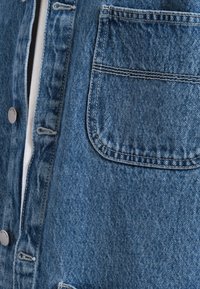 Giubbotto in denim di blu medio. Presenta chiusura con bottoni, taschino singolo sul petto, cuciture visibili e un aspetto in tessuto testurizzato.