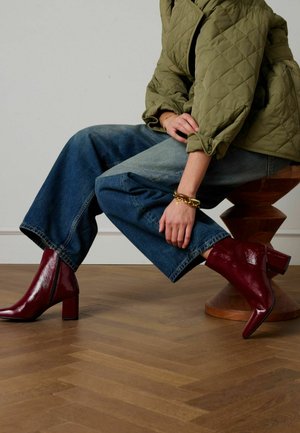 Bottes à la cheville en cuir verni bordeaux avec un talon carré, associées à un jean bleu denim à jambes larges et une veste matelassée vert olive. Socle en bois en arrière-plan.